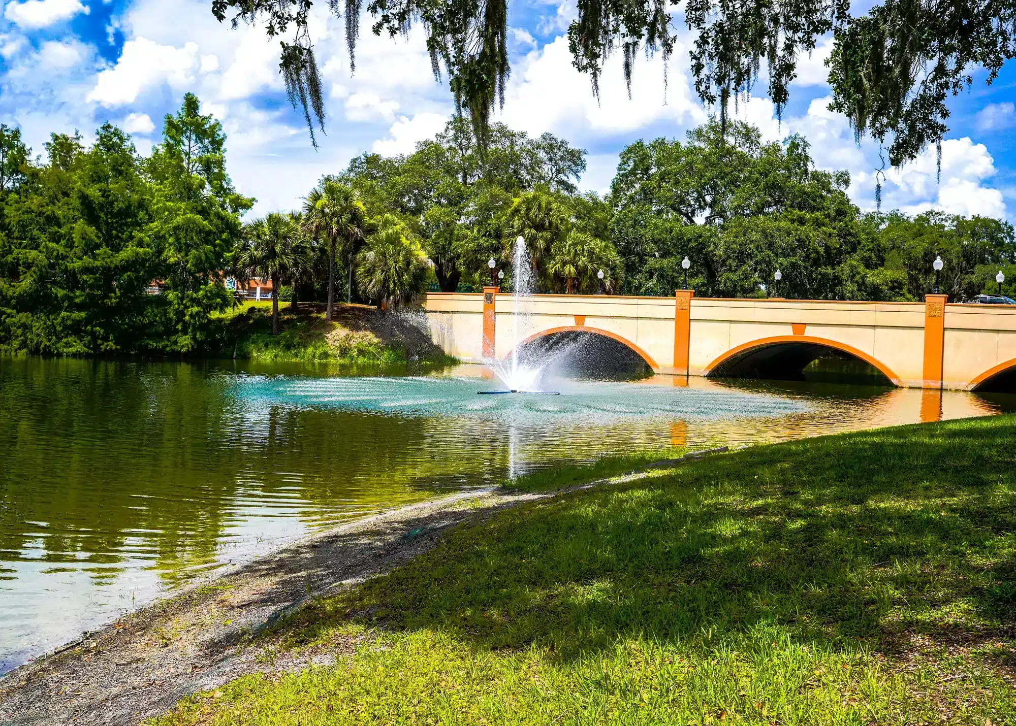 A calm park pond with a central fountain spraying water in front of a cream-and-orange three-arched bridge, framed by lush green trees, a grassy shoreline and a blue sky with hanging Spanish moss.