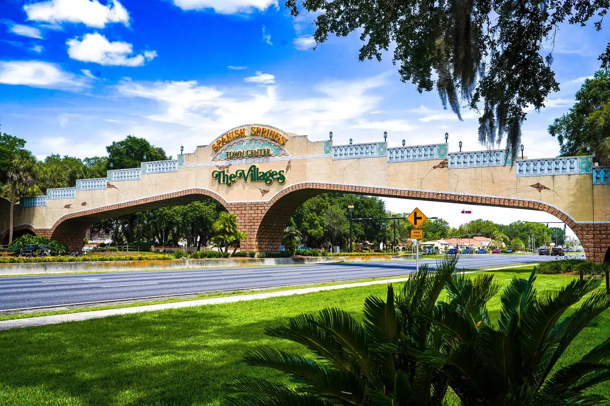 Decorative beige-and-brick arched gateway spanning a multi-lane road labeled “Spanish Springs Town Center” and “The Villages,” with palm trees, manicured green lawn in the foreground and a bright blue sky above.