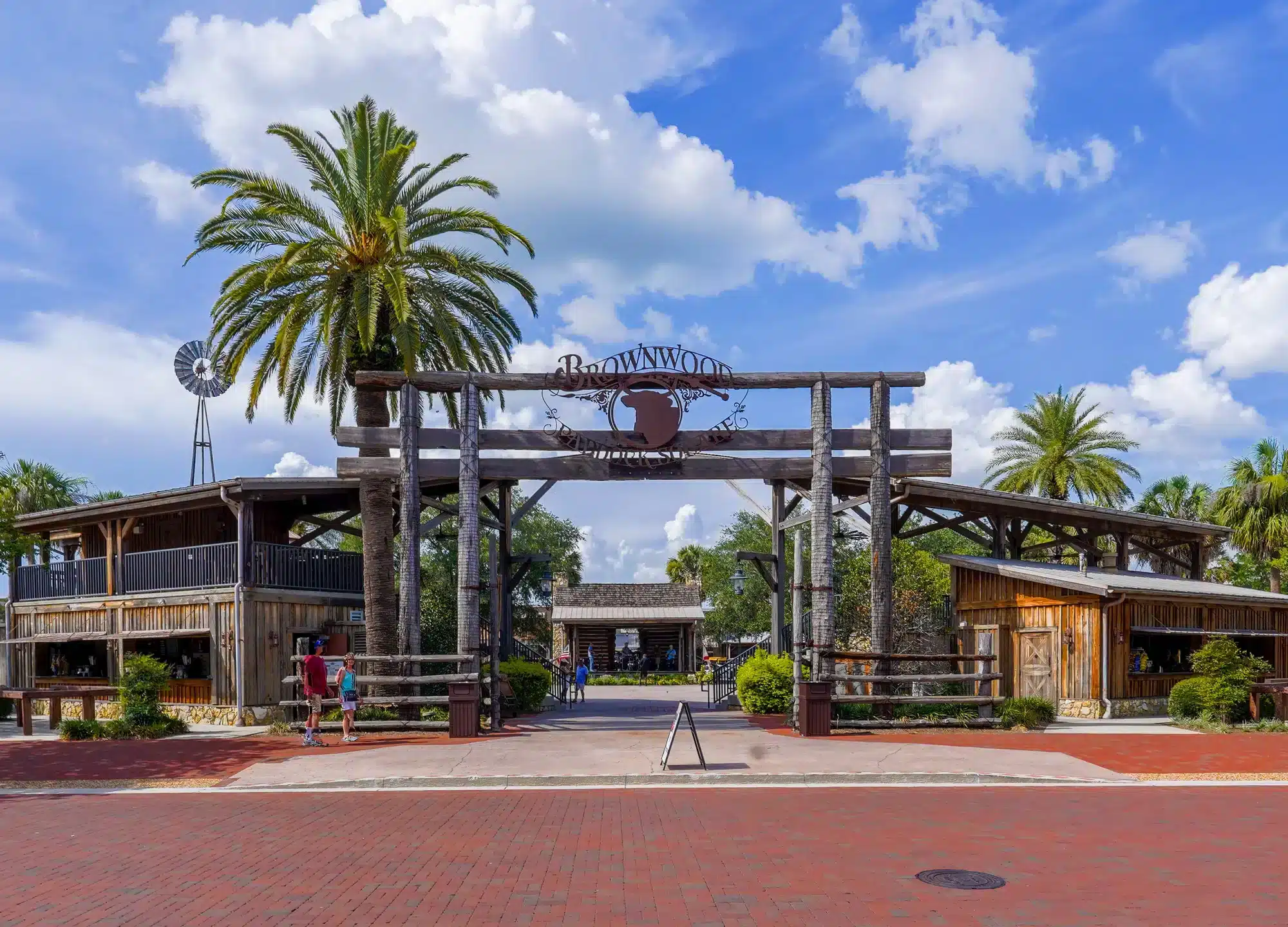 Rustic wooden entrance gate reading Brownwood framed by tall palm trees, flanked by two wood-clad buildings, with a brick plaza and a couple of pedestrians beneath a bright blue, cloud-dotted sky.