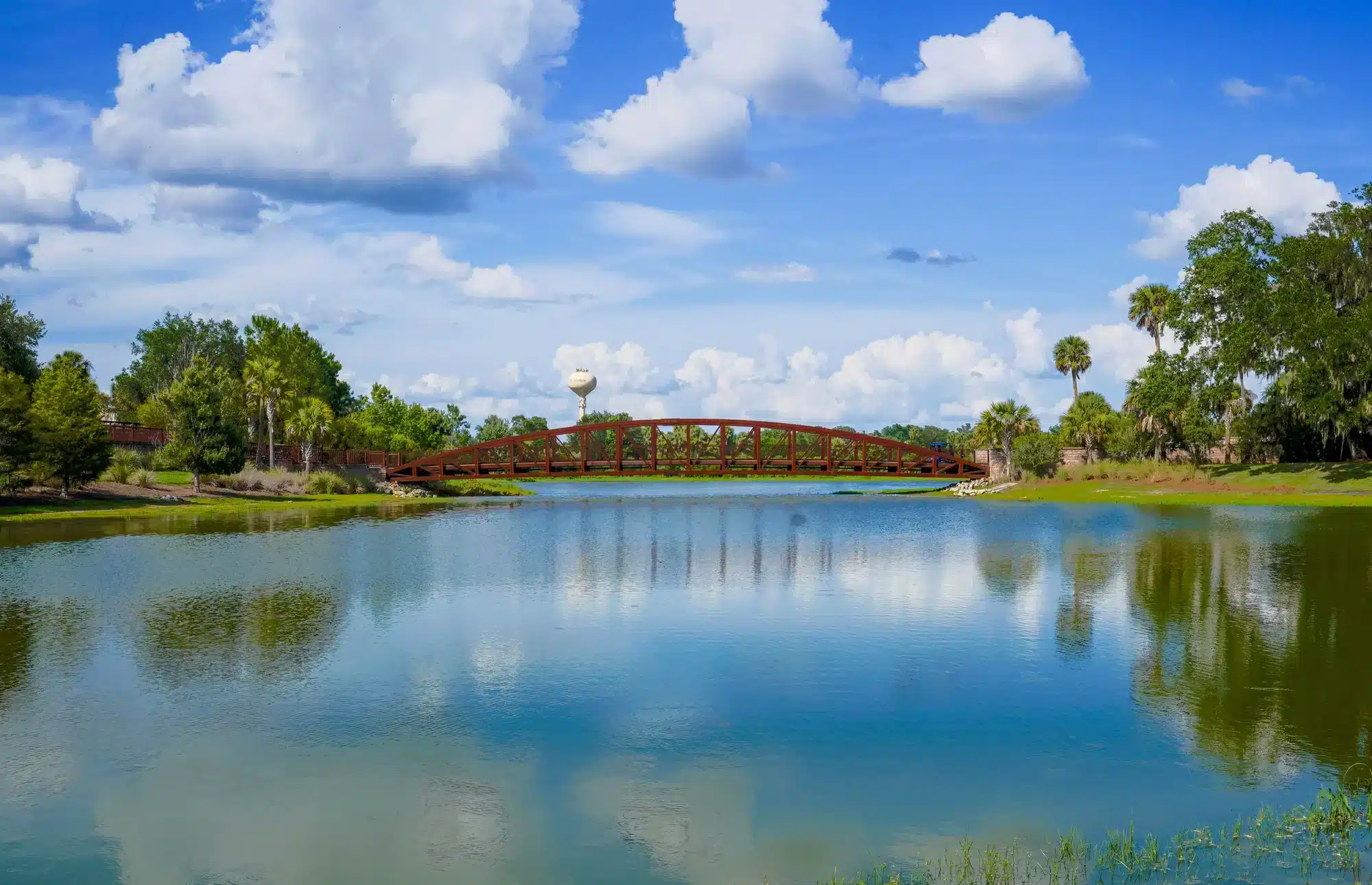 A low red pedestrian bridge crosses a calm lake that reflects fluffy white clouds and surrounding trees and palm trees, with a water tower visible in the distance under a bright blue sky.