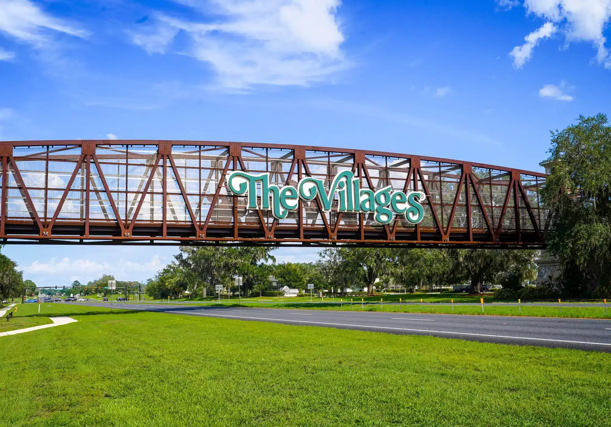 Rust-colored metal pedestrian bridge spans a roadway with a large green-script sign reading The Villages, set against blue sky and a grassy foreground.