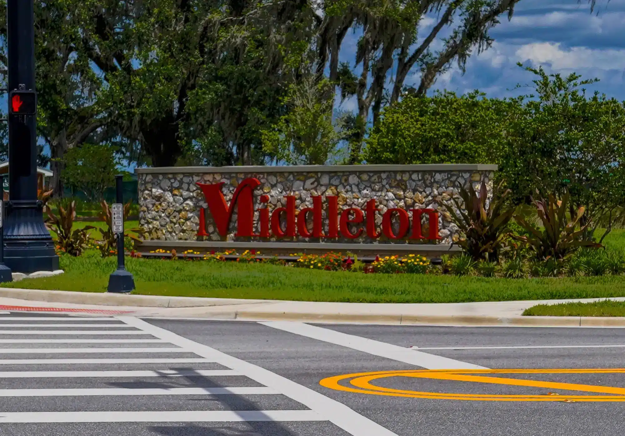 Stone entrance sign with large red letters reading Middleton, set in landscaped grass and flowers beside a pedestrian crosswalk and traffic signal, with trees and a cloudy sky behind.