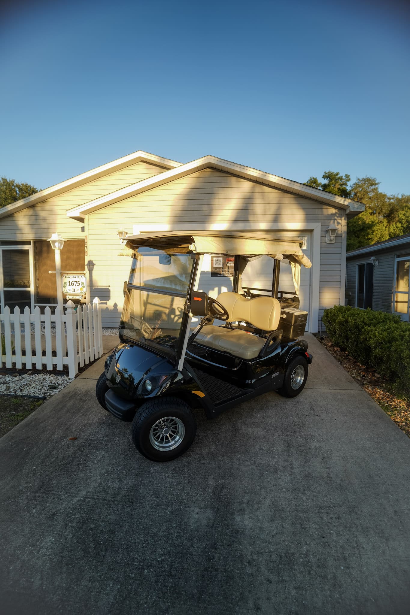 A black golf cart with beige seats parked in front of a house with light gray siding and a white picket fence, under a clear blue sky.