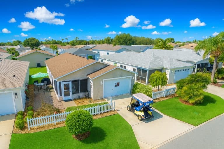 Single-story suburban house with white picket fence and a golf cart parked in the driveway, manicured lawns and blue sky.