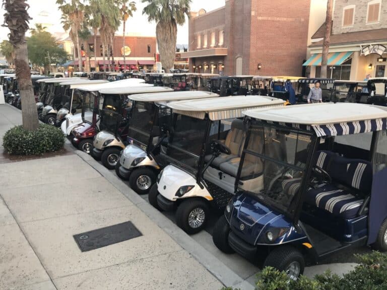 Row of parked golf carts with canopies lined along a downtown street in front of shops and palm trees.