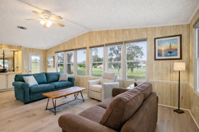 Living room with teal sofa, brown couch, beige armchair, wooden coffee table and row of windows overlooking a green lawn.