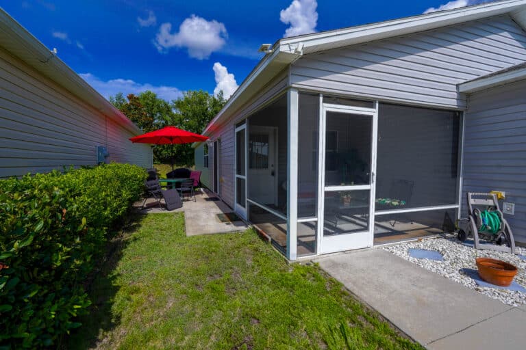 A screened porch and concrete patio sit between two siding houses, with a red umbrella, outdoor chairs, and a hose reel.