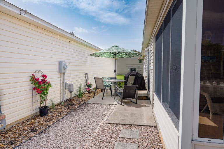 Narrow side patio with round table and chairs under a green umbrella, grill, potted flowers, gravel path and screened porch.