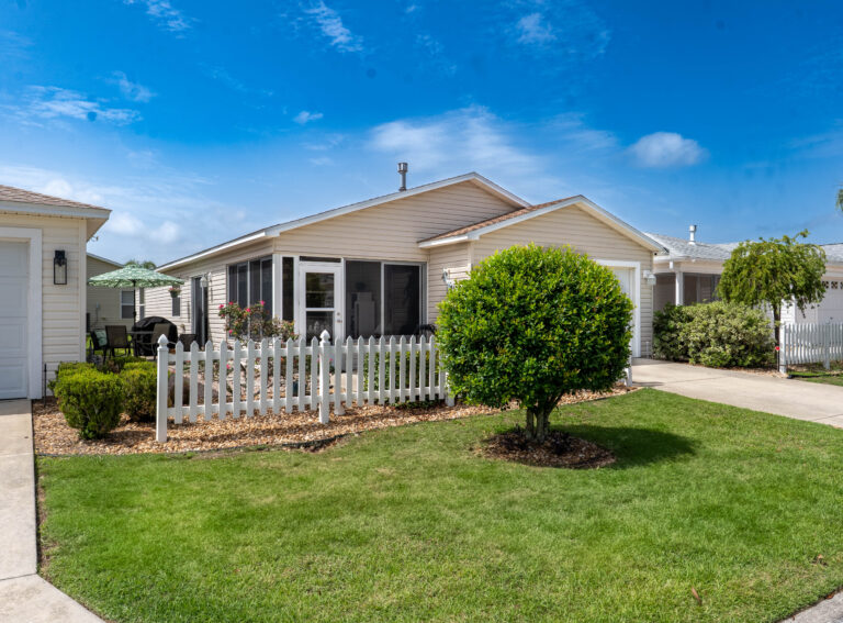 Single-story beige house with screened porch, white picket fence, round trimmed shrub and a manicured green lawn under a blue sky.