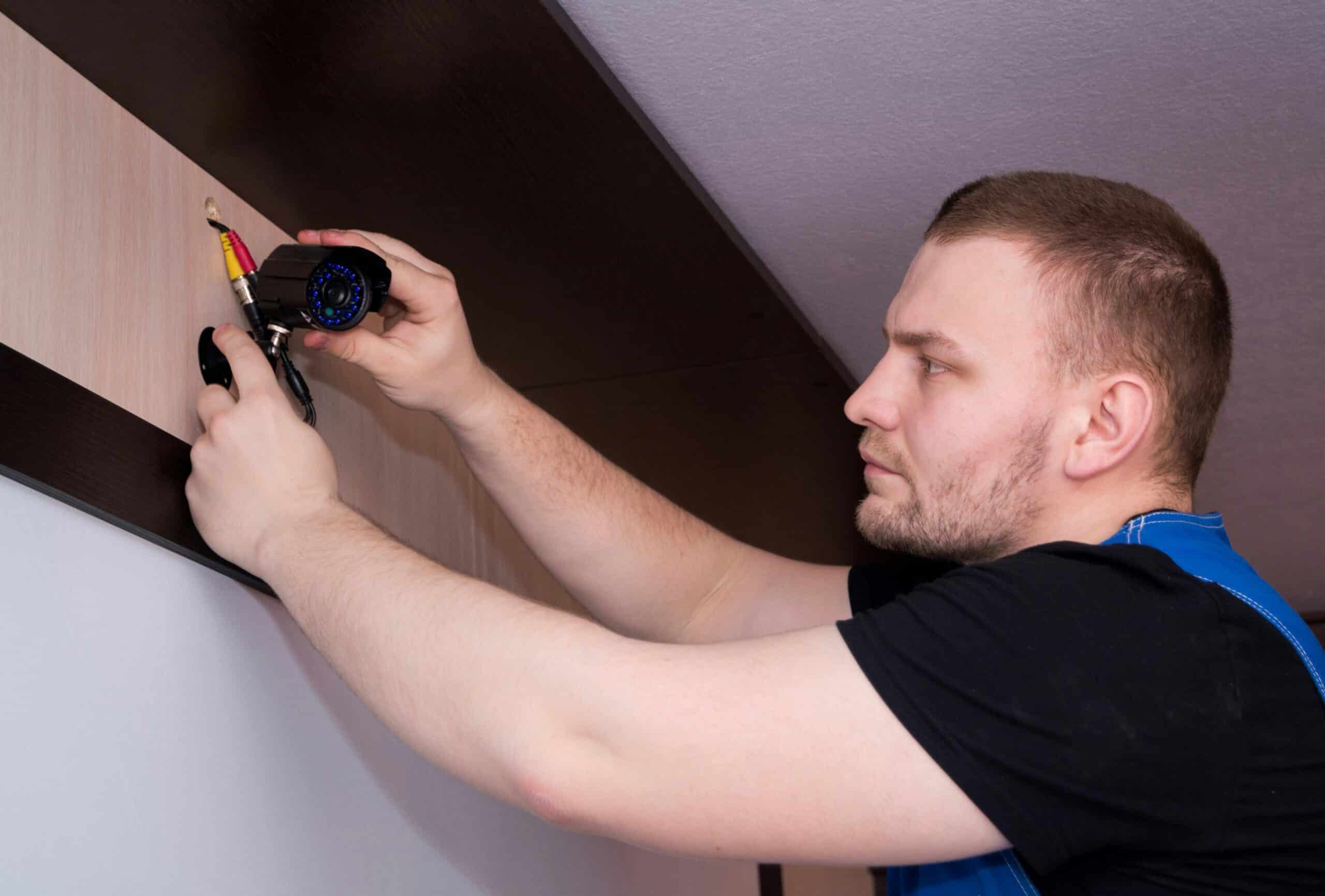 Man in blue overalls mounting a black security camera to a wooden ceiling beam while holding connector cables